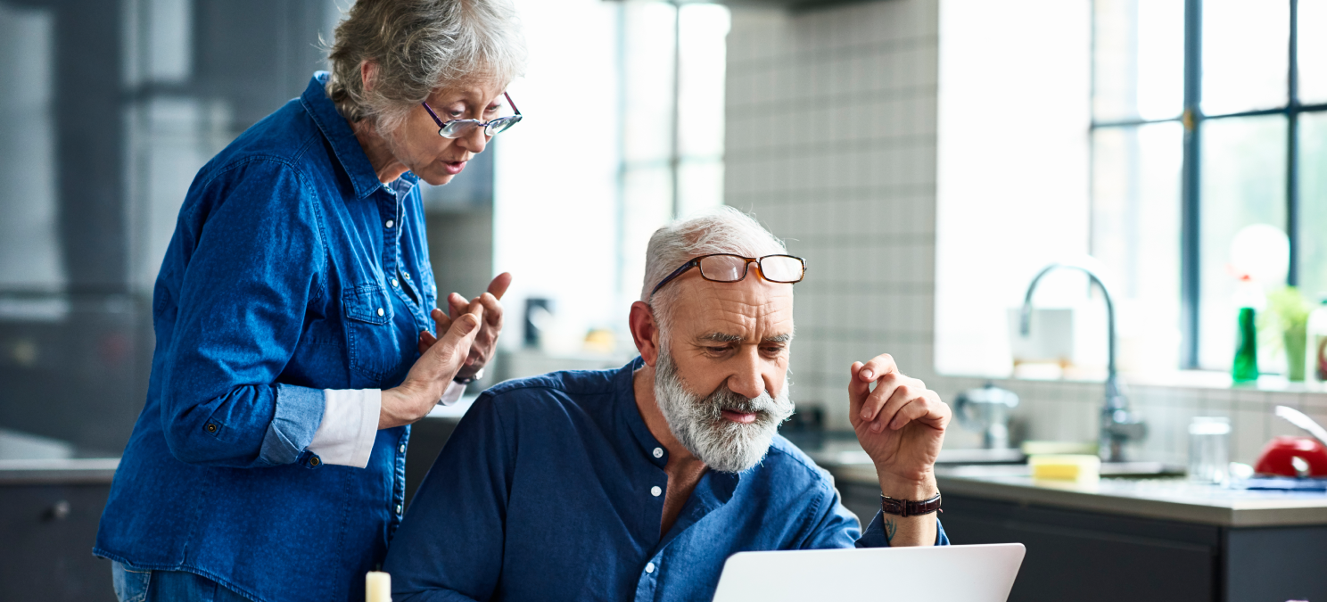 man sitting at table in front of laptop with woman standing behind.